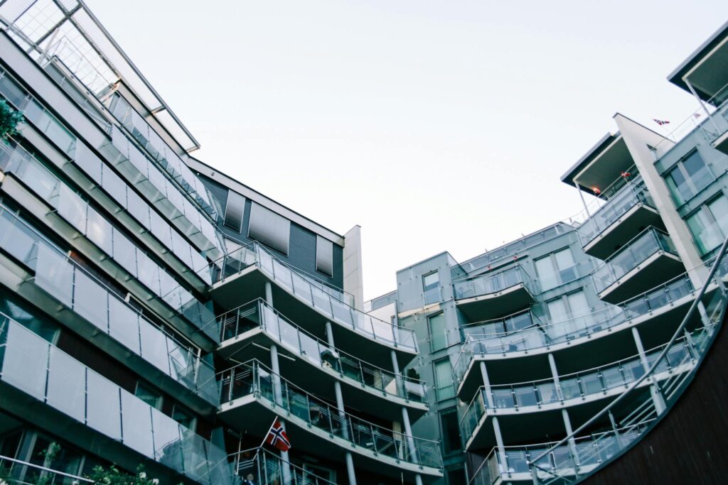 Low-angle view of a modern glass building featuring balconies and Norwegian flags.